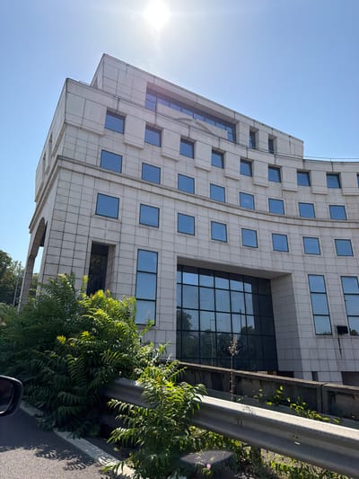 This media file captures an exterior view of a modern, multi-story building with a light-colored, tiled facade and numerous rectangular windows. The building's architecture features a stepped or angled design on the left side, creating a dynamic silhouette against a clear blue sky. The sun is visible in the upper left quadrant, casting a bright glare and suggesting it is daytime.
In the foreground and to the right of the building, lush green foliage, likely trees or bushes, is abundant. A metal guardrail runs parallel to the building, partially obscured by the greenery. The ground in the lower right appears to be asphalt, indicating a road or parking area. The orientation of the image is rotated, with the building extending diagonally from bottom left to top right.
There are no people visible in the frame. The overall scene is one of urban architecture combined with natural elements, suggesting a setting in or near a city. The bright sunlight and clear sky point to fair weather. There is no visible text in the image. Based on the provided location context, this scene is situated in Sèvres, France.