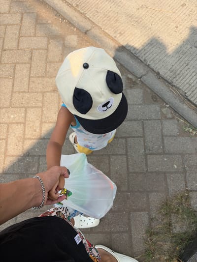 The image is an overhead shot of a person holding a child's hand as they walk on a paved path. The child is wearing a panda-themed hat and a light-colored shirt with a cartoon pattern. They are also holding a translucent plastic bag filled with green items. The person holding the child's hand is wearing a black top with a colorful, patterned lower garment and white clogs. A silver chain bracelet is visible on the person's wrist. The ground is made of interlocking rectangular pavers, with a strip of concrete and some dry grass to the left. The lighting suggests it is daytime, possibly late afternoon, given the long shadows. The scene appears to be outdoors in a public area, likely a sidewalk or walkway in Denasri Kulon, Indonesia. No discernible text is visible in the image.