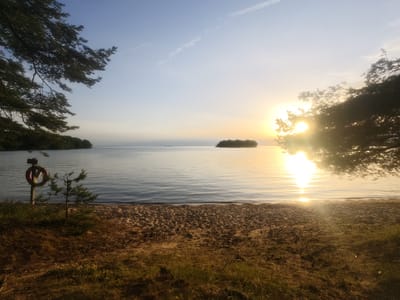 This image depicts a serene lakeside scene at sunrise in Motala, Sweden. The sun, low on the horizon, casts a warm golden glow across the calm water, creating a shimmering reflection. In the foreground, a sandy beach is bordered by dry grass and some scattered wildflowers, suggesting a dry climate or season. To the left, a wooden post supports a life preserver, a common safety object found near bodies of water. A small evergreen sapling grows near the post. The middle ground features the tranquil lake with a small, tree-covered island in the distance. The background is dominated by a clear sky, with a faint contrail from an airplane and a few wispy clouds. Overhanging pine branches frame the left and right sides of the image, adding a natural, woodland element to the lakeside setting. The overall mood is peaceful and tranquil, evoking the quiet beauty of early morning. The sun's low angle indicates it is either sunrise or sunset, but given the clear sky and gentle light, sunrise is more probable. No text is visible in the image.