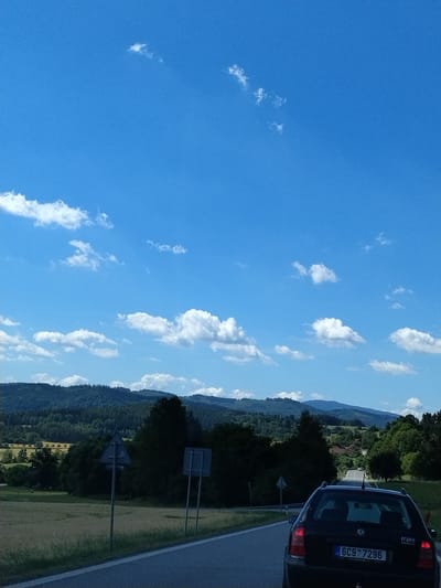 The image depicts a road trip scene on a sunny day, likely near Malenice, Czechia. A black Skoda Octavia is the primary subject in the foreground, its rear visible as it travels along a paved road. The car has a Czech license plate that reads "6C9 7296". The road curves slightly ahead, leading towards rolling hills covered in lush green forests. In the distance, more distant hills fade into a hazy blue sky dotted with scattered white clouds. The landscape also features fields and some scattered houses nestled within the trees. Two grey triangular traffic signs are visible along the roadside. The overall impression is one of a peaceful rural drive under a bright sky.