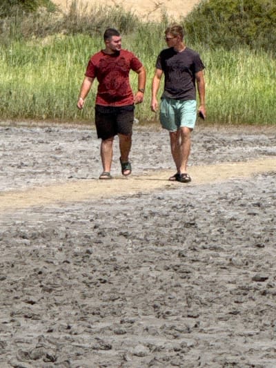 Two men are walking on a dry, cracked, muddy path. The man on the left is wearing a dark red t-shirt and black shorts. The man on the right is wearing a dark gray t-shirt and light blue shorts and is holding a dark object in his right hand. They are walking away from the camera, towards a background of green vegetation and light brown earth. The ground is covered in a textured pattern of dried mud and small rocks. The scene appears to be an outdoor, natural environment. The location is not specified as being in the city of Бурсоль, Russia.