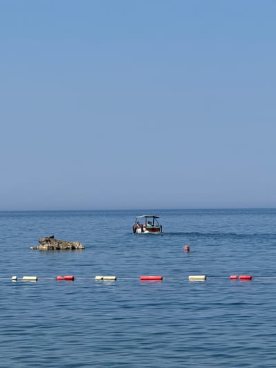 The image depicts a clear blue sky and calm, deep blue water, likely the Adriatic Sea, in Kamenovo, Montenegro. In the mid-ground, a small motorboat with a canopy is visible, carrying several people. To the left of the boat, a cluster of rocks protrudes from the water. In the foreground, a line of buoys, alternating between red and yellow, floats on the surface, marking a boundary in the water. A person is visible swimming in the water, to the right of the boat. The overall scene suggests a peaceful day at the beach or sea.