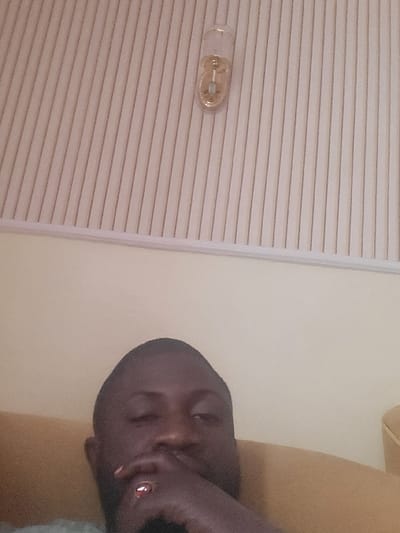 The image depicts an indoor scene in the city of Babawa, Nigeria. A dark-skinned male subject, visible from the nose upward, occupies the lower half of the frame. He has short hair and appears to be resting his chin on his right hand, which displays a reddish-brown ring on the ring finger. His eyes are partially closed. The subject is seated on a light brown or tan upholstered surface, possibly a sofa or armchair. The background consists of a plain light beige wall. Above the wall, a white panel with vertical striations extends across the upper portion of the frame. Mounted on this white panel, slightly right of center, is a transparent, capsule-shaped decorative object containing a golden interior and a central element.