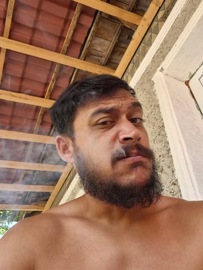 A close-up, low-angle selfie shows a shirtless man with dark hair and a full beard smoking. He is outdoors, with a wooden roof structure and a tiled roof visible in the upper left background. To the right, a textured wall with decorative tiling and white trim is present. The man's face is turned slightly towards the camera, with smoke emanating from his mouth. The background suggests a rustic or rural setting, possibly in Drianovets, Bulgaria, given the traditional roofing materials and exterior wall construction. The lighting indicates it is daytime, with sunlight illuminating the scene.