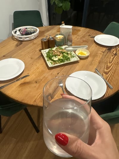This is a medium shot, eye-level, indoor photograph of a round wooden dining table set for a meal. The image is taken from a first-person perspective, with a hand holding a clear glass of water in the foreground. The table is made of wood with visible grain and has four dark green upholstered chairs surrounding it.
On the table, from left to right, is a white decorative bowl filled with various items, possibly personal effects like keys, masks, and a wallet. Next to it are a salt and pepper shaker set, followed by a lit candle and a small bottle. In the center of the table, a rectangular white dish contains a salad with greens, nuts, and possibly cheese or fruit. To the right of the salad is a small white dish with three wedges of lemon. Further to the right are two white plates, one with a knife placed to its right and the other slightly out of focus.
The overall environment suggests a home dining setting, possibly in Bonville, Australia, as inferred from the prompt. The lighting is warm and ambient, likely from overhead or nearby artificial light sources. The background is dark and out of focus, but a hint of a plant and dark furniture can be discerned, suggesting an indoor, domestic space. The hand in the foreground has red nail polish.