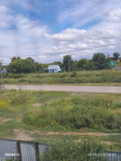 The image captures a rural or suburban landscape under a cloudy blue sky. A paved road runs horizontally across the mid-section of the frame. On the far side of the road, there is a densely wooded area with a light blue house featuring a grey roof situated in the clearing. The foreground consists of a grassy slope with some dirt paths and a corrugated metal fence. The overall impression is of a calm, natural setting. The text "REDMI 15" and "28.06.2026 13:47" are watermarks on the image.