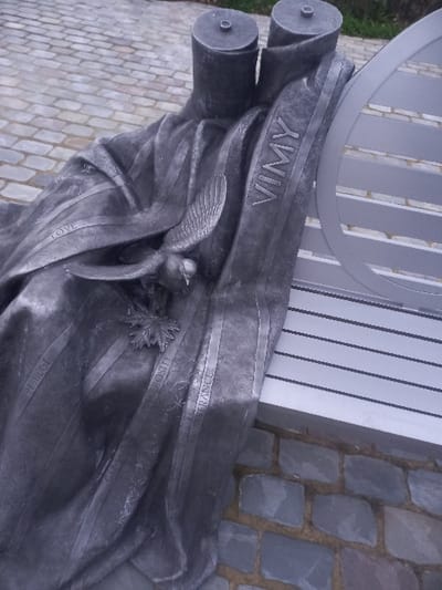 An abstract sculpture, likely metallic, features "VIMY" prominently on a banner-like segment. Below, a stylized bird with outstretched wings perches near a floral design. Other words, including "LOVE," "RESPECT," "GENEROSITY," and "FRANCE," are inscribed along decorative bands. The sculpture sits on a cobbled ground next to a modern metal bench. The overall composition is solemn and commemorative in tone.