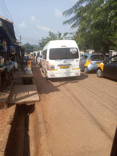 This image depicts a congested, unpaved street scene in Konongo, Ghana. The perspective is from slightly above ground level, looking down a reddish-brown dirt road with visible tire tracks.
In the foreground, a large, open concrete drainage ditch runs parallel to the left side of the road. A single flat concrete slab partially covers a section of this ditch. To the left of the road, a series of makeshift market stalls or shops with corrugated metal roofs are visible. Various items, including bags, plastic containers, and other goods, are displayed on tables in front of these structures. One person is seated under a stall, obscured, facing towards the road. Overhead, power lines or communication wires are strung across the scene.
The road itself is occupied by a line of vehicles. The most prominent is a white Toyota Hiace-style passenger van, observed from the rear, featuring a Ghanaian license plate "AS 8912-23" on a yellow plate. The rear window displays a sticker with "OCCUPY TILL I COME" and another depicting a "HOLY BIBLE" with a cross. A red circular sticker with the number "2" is also present. The van's bumper has white and yellow reflective tape. Directly behind this van, a queue of similar white passenger vans extends into the distance, suggesting traffic or a waiting area.
On the right side of the road, a light blue or silver small hatchback, possibly a Chevrolet Spark, with yellow lower body markings indicative of a Ghanaian taxi, is stationary or moving slowly. Behind it, a dark-colored sedan, also marked with yellow for taxi service, is visible. The right side of the road is bordered by a line of large trees with dense green foliage, with a low concrete wall or barrier partially visible beneath them.
The sky is a clear blue with some scattered white clouds, and strong sunlight casts distinct shadows, indicating daytime. No direct interactions between individuals are discernible. The scene illustrates typical commercial and transit activity in a Ghanaian urban environment.