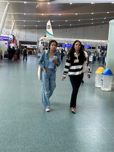 In Tambon Mai Khao, Thailand, two young women walk through a modern airport terminal. The woman on the left, wearing a denim jacket over a light blue tank top and wide-leg jeans, holds a clear plastic cup with a dark beverage in her right hand. A canvas tote bag hangs from her left shoulder. The woman on the right, with long dark hair, is dressed in a black and white striped long-sleeved shirt and black trousers, wearing sandals. She holds a smartphone in her right hand.

The environment is a well-lit airport concourse with a speckled grey-blue floor. Overhead, a slatted ceiling with integrated lighting is visible. In the background, airport check-in counters with signs displaying "IndiGo" and "B01," "B02," etc., are prominent. A "STARBUCKS COFFEE" sign is also visible, indicating commercial establishments within the terminal. Several other people are present in the background, some walking and others near the check-in counters, suggesting an active travel hub. Two large cylindrical trash receptacles, one yellow and one blue, are positioned to the right of the two women. The scene captures a moment of transit within a bustling airport.