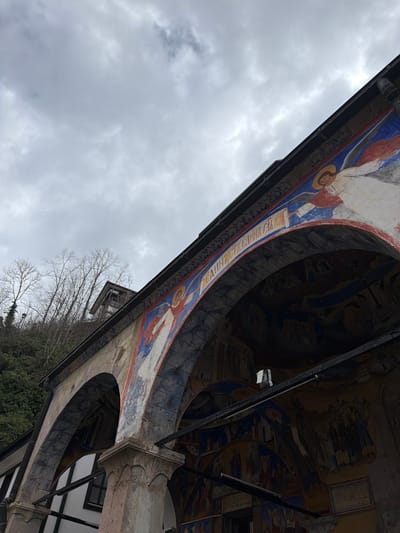 The image presents a low-angle view of an ornate architectural structure, possibly an arcade or portico, in Kriva Palanka, North Macedonia. The structure features arches decorated with paintings of figures and patterns in red, blue, white, and gold hues. Some inscriptions are visible on the painted arches. The ceiling beneath the structure appears to be covered in frescoes or detailed murals. Exterior architectural details, including pillars and window frames, are visible. The background consists of trees with bare branches, suggesting a season with less foliage, and an overcast sky. The image is taken from a position looking upwards towards the structure and the sky.