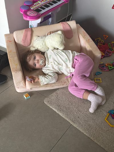 A child lies on a tan, cushioned child's seat with arms, indoors, oriented with her head to the bottom of the frame. She is dressed in a white button-up pajama top with green bear prints, pink pants, and white socks. She holds a white teddy bear and a pacifier in her left hand, and has a pacifier in her mouth. A pink toy keyboard sits to her left. Magnetic toy pieces surround the seat, on a neutral colored rug. Part of the floor is tiled, gray. A block rests at the lower right corner of the seat. Parts of a person's shoes are visible at the bottom of the frame. This scene appears to be within a home. Harish, Israel is specified as the city.
