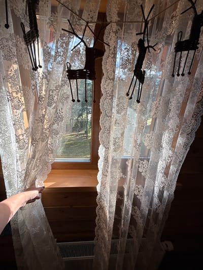 The image shows a close-up view of a lace curtain covering a window. The curtain is white and has a floral pattern. It is draped and slightly transparent, allowing sunlight to filter through, creating a soft, diffused light. The window looks out onto a scene with greenery, suggesting an outdoor setting, possibly a garden or yard.

In the foreground, a hand with dark nail polish is reaching out and holding onto the edge of the curtain, pulling it slightly. This action suggests movement or adjustment of the curtain. The lighting indicates it is daytime, with strong sunlight casting shadows and highlighting the texture of the lace.

There are also decorative metal elements attached to the curtain, resembling stylized deer or reindeers, with thin legs and antlers. These add a whimsical touch to the scene. In the background, a wooden wall is visible, and to the right, a white vent or air conditioner unit is partially obscured by the curtain. The overall impression is one of domesticity and a peaceful, sunlit moment. No text is visible in the image. The location context is Сосновка, Russia.
