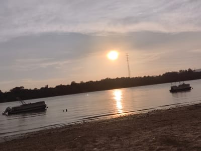 This media file captures a serene lakeside scene at Dunakeszi, Hungary, during what appears to be sunset. The sky is a soft gradient of pale orange and gray, with wispy clouds indicating a calm weather condition. The sun, a bright orb, is visible in the upper center of the frame, casting a shimmering reflection on the water.
The main subjects are two pontoon boats on the lake. The boat on the left is closer to the shore and features an outboard motor. There are also two figures visible in the water near this boat, suggesting some form of recreation or swimming. The boat on the right is further out in the lake and appears to be anchored or drifting.
The foreground consists of a sandy beach with some scattered debris, including a rope. The background is a dark silhouette of trees lining the opposite bank of the lake, with a tall electricity pylon also prominent in the distance. The overall mood is tranquil and peaceful, indicative of a late afternoon or early evening on the water. There is no visible text in the image.