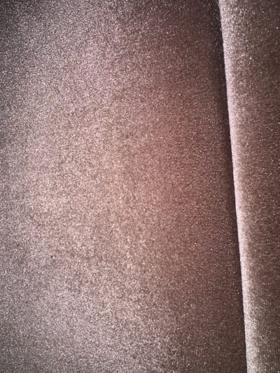 The image displays a close-up, textured view of fabric. The fabric appears to be a light brown or taupe color with a subtle shimmer, likely due to metallic threads or a glittery finish. The texture is fine and somewhat velvety. A fold or crease in the fabric is visible in the upper left portion of the image, creating a diagonal line that divides the frame. The lighting is somewhat dim, with reflections highlighting the glittery particles within the fabric. There are no discernible subjects, activities, or text in the image. The location context of Gülnar, Turkey, does not provide any visual cues for interpretation within the image itself.