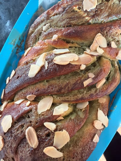The image depicts a baked loaf of twisted bread in a blue baking pan. The bread is golden brown on the exterior, with a green-tinged interior. It is adorned with sliced almonds. The bread appears to be sitting on a counter. The location is not discernible from the image.