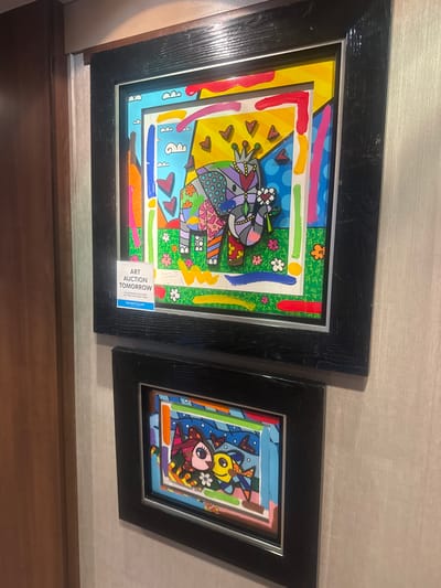 The image displays two framed pieces of art hanging on a wall with a textured, light-colored finish. The frames are black and appear to be made of wood. The left frame contains a white informational card that reads "ART AUCTION TOMORROW" with additional details about the auction location and time.
The artwork itself is brightly colored and features a whimsical, cartoonish style. The upper artwork depicts an elephant wearing a crown and holding a flower. The lower artwork features a stylized depiction of a girl with a witch's hat and a yellow fish. The art appears to be in the style of Romero Britto, known for his vibrant use of color and bold patterns.
The location is identified as Motril, Spain, and the context suggests this is a display within a gallery or event space related to an art auction.