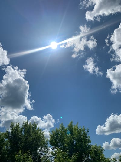 This media file depicts a bright, sunny sky with scattered white clouds, as viewed from below. The sun is a prominent element, positioned in the upper left quadrant, casting a strong, white beam of light that streaks diagonally across the image. The sky is a clear, vibrant blue. On the right side of the frame, the lush green foliage of a tree canopy is visible, extending into the sky. There are no people or specific objects that indicate a particular event or activity. The scene suggests a daytime setting, likely mid-morning to early afternoon, given the intensity of the sunlight. The weather appears fair and clear, with the presence of cumulus clouds indicating a typical summer day. Location cues are limited to the suggestion of trees and a sky that could be found in many outdoor settings. No text is visible in the image.