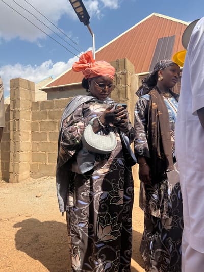 Two women stand side-by-side in Rigasa, Nigeria. Both wear brown floral print dresses and head coverings. The woman in the foreground has a peach-colored head wrap, sunglasses, henna tattoos on her hands, and carries a light gray purse. She holds a smartphone in her left hand. The woman behind her wears a patterned dark head wrap. They are outside in an area with brick structures and a corrugated metal roof. A utility pole and wires are visible against a blue sky with clouds. The ground appears to be dirt or sand.