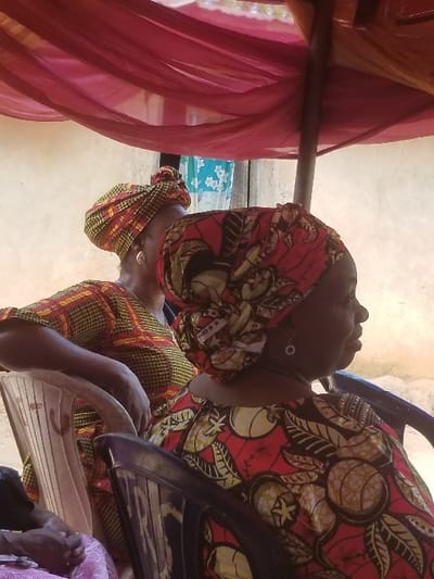 An outdoor scene in Ochadam, Nigeria, features two individuals seated beneath a red sheer fabric canopy. In the foreground right, a woman is seen in profile, wearing a red dress with yellow and green fruit and leaf patterns, a matching elaborate head wrap, and a circular silver earring. She is seated in a dark plastic chair. To her left and slightly behind, a second woman is partially visible, facing left, wearing a yellow and red plaid-patterned dress and a matching head wrap. She is seated in a light-colored plastic chair, with her left hand resting on the chair's back. The background consists of a light-colored building wall, with a partial view of a blue floral-patterned fabric further left. A dark support pole is visible under the canopy.