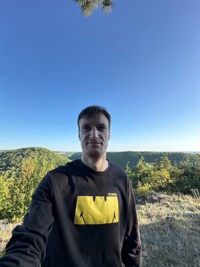A man stands for a selfie on a hillside in Pryvorottia Druhe, Ukraine. He is wearing a black sweatshirt with a yellow smiley face graphic and the text "THE ORIGINAL" above it. The background is a lush, green landscape with rolling hills and a clear blue sky. The sun appears to be setting, casting a warm glow over the scene. The man is looking directly at the camera with a neutral expression.