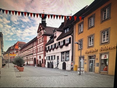 The image shows a street scene in Ellwangen, Germany, featuring a row of historic buildings decorated with festive triangular flags in red, black, and blue strung across the street. The buildings are of various architectural styles, with facades of yellow, white, and red brick. One building on the right has a prominent sign that reads "Apotheke Goldener," indicating a pharmacy. A street lamp is visible in front of the pharmacy. The street is paved with cobblestones, and large potted plants are placed along the sidewalk. Several people are walking on the street, and one person is seen entering the pharmacy. The sky is cloudy, suggesting an overcast day.