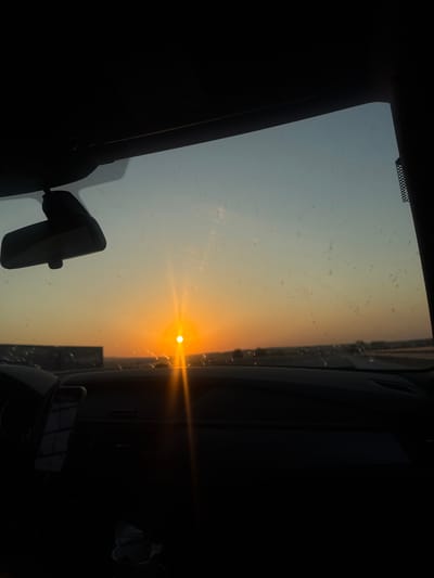 The image, taken from inside a vehicle, captures a sunset over what appears to be an open landscape, potentially near Cherna Gora, Bulgaria. The sun, a bright orb of orange, is low on the horizon, casting a warm glow across the sky. The sky transitions from a vibrant orange near the sun to a pale blue at the top of the frame. The windshield of the car is visible, with some smudges and streaks that are illuminated by the sunset. A rearview mirror is mounted on the windshield. The dashboard of the car is in the foreground, largely in shadow, with a phone mounted on the passenger side. The overall impression is one of a scenic drive during twilight.