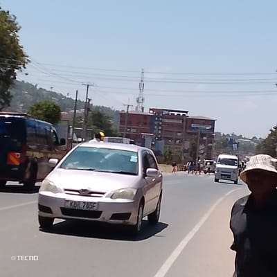 A daytime street scene in Machakos Central ward, Kenya, depicts a multi-lane paved road under a clear blue sky. In the immediate foreground on the right, the head and upper torso of an individual wearing a patterned bucket hat and a dark top are partially visible. A light-colored (champagne/silver) Toyota Corolla sedan, bearing Kenyan license plate KBR 785F and equipped with a yellow roof-mounted taxi sign, occupies the central mid-ground, heading left relative to the viewer. To the left of the Corolla, a dark blue van or minibus with a red-orange reflector on its rear is partially visible. Further down the road, a white van and several indistinct pedestrian figures are present. The background features several multi-story buildings, notably one with prominent red and dark blue sections, along with a tall cellular tower and utility poles carrying overhead lines. Hilly terrain with sparse vegetation is visible in the distant background. Bright sunlight illuminates the scene, casting shadows. A "©TECNO" watermark is present in the lower-left corner.