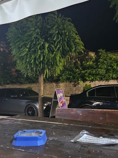 The image depicts a nighttime scene on a wooden table in front of a building. In the foreground, a blue ashtray sits on the table. Behind the table, a street is visible with two parked cars, one dark and one gray, a tree, and a stone wall. A small advertising stand with the "Milka" logo is positioned near the cars. The background shows greenery and parts of buildings. The location is identified as Byala, Bulgaria, suggesting an outdoor seating area of a cafe or restaurant.