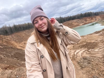 A woman stands in the foreground, wearing a beige trench coat and a mauve beanie. She has long brown hair and is smiling slightly.
The background shows a large excavated area with a lake at the back. The terrain is mostly brown earth. Trees are visible along the periphery of the excavated area. The sky is overcast with dark clouds.
The location is Ivlevo, Russia.