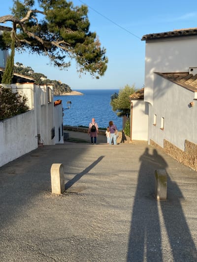 The image captures a street in Calella De Palafrugell, Spain, with a view towards the Mediterranean Sea. Two people, an adult wearing a purple shirt and jeans, and a child, are walking down the street towards the sea. Buildings line both sides of the street, characterized by white facades, tile roofs, and rectangular windows. The street itself is paved and features concrete blocks protruding from the side. Shadows cast by the structures and possibly trees create an interesting contrast on the street surface. In the background, the blue sea is visible along with a small, rocky landmass. A large tree is prominent on the left side of the frame, contributing to the overall atmosphere of the scene.