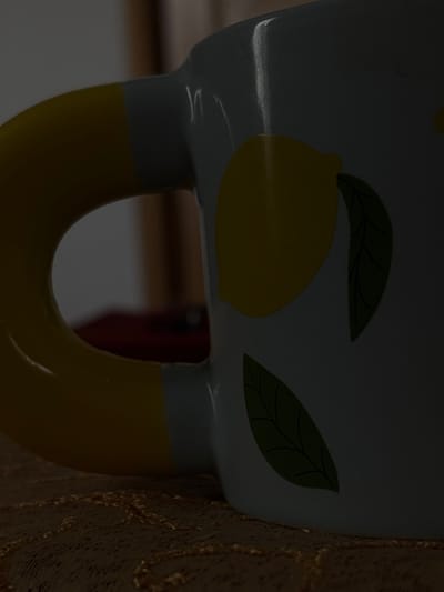 The scene is an interior, located in Griboyedov, Armenia. A white ceramic mug with a prominent yellow, C-shaped handle occupies the majority of the frame, positioned on a textured, light brown or golden surface. The mug's exterior features yellow lemon designs accompanied by dark green leaves. Illumination is dim, casting the mug and environment in low light, with subtle reflections visible on the mug's glossy surface. Through the mug's handle, a blurred, dark background is visible, containing indistinct shapes suggestive of indoor furniture or structures, including a faint reddish object. No individuals are present, and no actions or interactions are depicted.