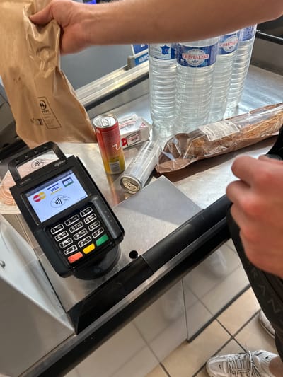 The image depicts a point-of-sale transaction at a supermarket checkout in Alès, France. A hand is holding a brown paper bag, likely containing purchased items, and is in the process of placing it on the counter. Several bottles of Cristaline water are lined up behind the bag. A baguette in plastic packaging lies on the counter next to a can of what appears to be Red Bull and another metal can. A payment terminal, an Ingenico Lane/5000, is positioned prominently on the counter, displaying "Visa" and "Mastercard" logos, indicating it accepts card payments. The screen also shows "Infomil" and a date of "20/06/2026 17:43". The terminal has a keypad with numerical buttons and function keys. A person's hand is reaching towards the payment terminal. The floor is tiled in a beige color. The scene is illuminated by overhead artificial lighting. The environment suggests a typical retail setting.
