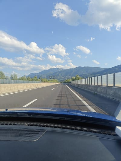 The image depicts a view from the driver's perspective in a vehicle traveling on a multi-lane highway. The road ahead is paved asphalt with white lane markings. On either side of the highway are concrete barriers, with transparent sound barriers installed on top of the right-side barrier. Beyond the barriers, trees and green foliage are visible. In the background, a range of mountains under a blue sky with scattered white clouds is prominent. The vehicle's dashboard and part of the windshield frame are visible in the foreground. The scene appears to be an urban or suburban area, possibly on the outskirts of Inzersdorf im Kremstal, Austria, given the presence of sound barriers common along highways near populated areas. There are no people or active events visible in the image.