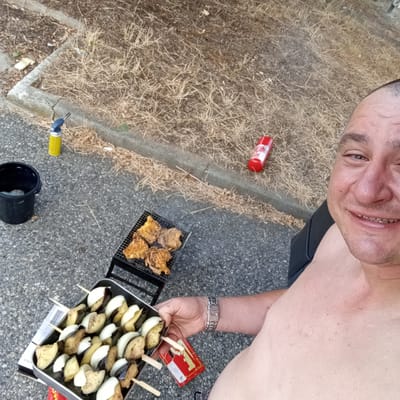 The image shows a close-up selfie of a man taken outdoors. He is smiling and appears to be shirtless. In the background, there is a small charcoal grill with pieces of food, likely chicken, cooking on it. To the left of the grill is a black bucket. Further to the left, a yellow butane torch is visible, suggesting the grill is being lit or maintained. In front of the grill, a silver tray holds skewers of what appear to be eggplant and onion, prepared for grilling. A red can, possibly a beverage or fire starter, is lying on the dry, grassy ground near a concrete curb. The setting appears to be a casual outdoor gathering, possibly a picnic or backyard barbecue. The lighting suggests it is daytime, with no strong indication of weather conditions. The man's expression is joyful and relaxed, hinting at a pleasant and enjoyable activity. A red cigarette pack with white lettering is visible near the tray of skewers, with the brand name "Budweiser" partially obscured. The man is wearing a silver watch on his left wrist. The overall scene conveys a sense of simple pleasure and outdoor enjoyment.