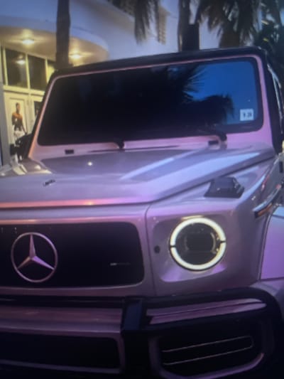 The image shows a close-up of the front of a light pink Mercedes-Benz G-Class SUV. The vehicle has a prominent Mercedes-Benz star logo on the grille and round headlights with distinctive LED daytime running lights. The body of the car appears to be a metallic or iridescent pink, reflecting ambient light and colors. The windshield is dark and reflective, showing the silhouette of palm trees and a blue sky. A small white sticker with black lettering and numbers is visible on the lower right corner of the windshield. In the background, a building with large windows and doorways is visible, with what appears to be a person standing near one of the entrances. Palm trees are also present in the background, suggesting a warm climate or a location with a tropical or Mediterranean influence. There are also exterior lights visible on the building. The image quality is somewhat low, with some blurriness and pixelation. The scene appears to be in an urban or resort-like setting, possibly during twilight or evening due to the dark sky and artificial lighting. The provided location of Kołobrzeg, Poland, does not align with the visual cues of palm trees and what appears to be a warm climate, suggesting a discrepancy in the provided information.