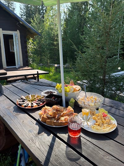 The image depicts an outdoor picnic scene set on a rustic wooden table in the city of Kuliki, Russia. A dark brown wooden cabin with an open door is visible in the background, surrounded by lush green trees and foliage, suggesting a wooded or natural setting. The table is laden with various food items, including a platter of olives and rolled meats, a bowl of mixed fruits, a salad in a glass bowl, a plate of cut pizza, and a cheese platter with various cheeses and grapes. A glass of red beverage and two small glasses of orange juice are also present, along with small containers of nuts and olives. An open green umbrella provides shade over a portion of the table. The overall atmosphere is one of a leisurely outdoor gathering.