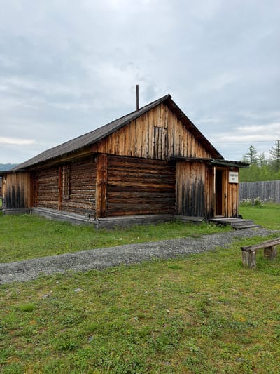The image shows a wooden building with a dark metal roof in Ust'-Pyzas, Russia. The building is constructed from logs and has a pitched roof. There is a small, open doorway on the right side of the building with a wooden porch and steps leading up to it. A sign with Cyrillic writing is visible above the doorway. The building is surrounded by green grass and a gravel path. In the background, there is a wooden fence and trees. A rustic wooden bench is situated in the foreground to the right. The sky is overcast.