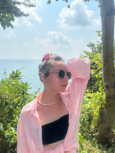 A woman is lying in an outdoor setting in Ao Hin Khok, Thailand. She is wearing a black bikini top and a light pink, unbuttoned shirt. The woman is also wearing sunglasses and a pearl necklace. There is a pink flower in her hair. The background shows a blue sky with scattered clouds and the ocean, along with lush green vegetation. A tree branch is positioned across the top of the frame.