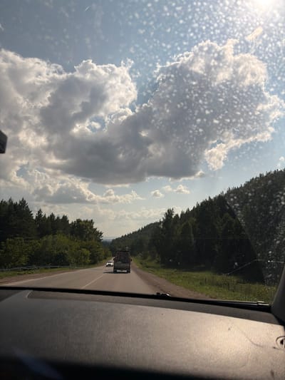 The image depicts a view from inside a vehicle traveling on a road. The road is paved and curves gently to the left. On the right side of the road, there is a dense forest of green trees extending up a hillside. On the left side of the road, there is a lighter green forest and a guardrail. In the distance, a white car is visible on the road, followed by a truck with a large cargo area. The sky is blue with scattered white clouds, and the sun appears to be shining, casting bright light. The foreground of the image shows the dashboard of the vehicle, which is dark and textured. Small water droplets or debris are visible on the windshield, creating a speckled effect. The scene appears to be a rural or suburban landscape in Старые Туймазы, Russia, possibly during daytime. There are no people clearly visible in the image.