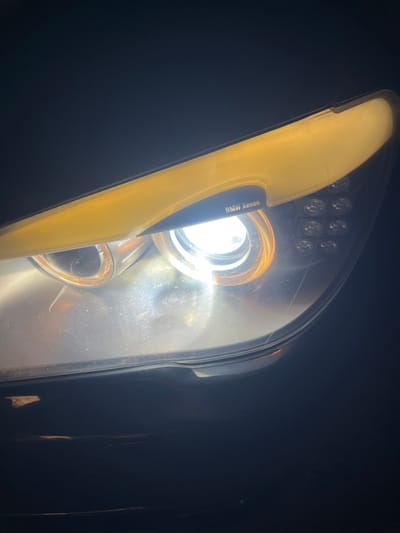 The scene depicts a close-up, angled view of the illuminated headlight of a BMW vehicle, located in Zlatovrah, Bulgaria. The headlight assembly features a central, bright white Xenon projector light surrounded by two concentric amber "angel eyes" rings. Above the main light, an elongated, illuminated yellow strip forms the upper edge of the headlight housing. To the right of the central projector, a cluster of nine smaller, circular, unlit lenses is visible. The words "BMW Xenon" are embossed in white text on a dark, triangular section above the main light source. The clear plastic lens covering the headlight exhibits surface dust or minor imperfections. The surrounding environment is dark, with the lower portion of the image revealing a glimpse of the vehicle's dark bodywork. The headlight is active, casting a strong light into the dark surroundings.