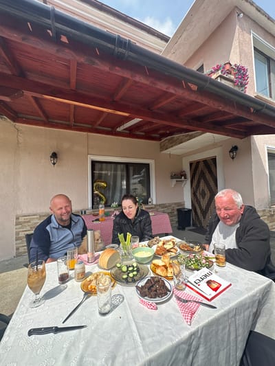 The image captures a family gathering outdoors in Vinarovo, Bulgaria. Three individuals are seated around a table laden with food and drinks. An elderly man with white hair sits at one end, wearing a dark jacket over a white shirt. A woman in a black jacket sits along the table, and a bald man in a blue and grey jacket sits at the other end, smiling. The table is covered with a white tablecloth patterned with a floral design, and food items include bread, meats, salads, and various other dishes. A box of Merci chocolates is also visible. Glasses and silverware are scattered around the table. In the background, the side of a house is visible with a door and window. A decorative rug hangs above the door. A black drain pipe runs along the edge of the house. A window box filled with flowers sits on the building behind the family. Balloons are visible in the window. A dog is lying on the table between the woman and the bald man. The scene is brightly lit, suggesting a sunny day.
