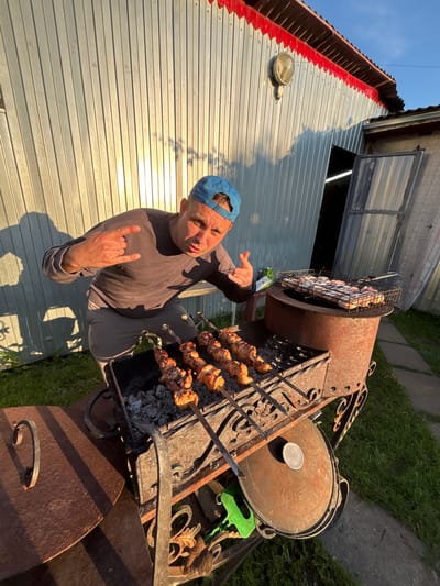 The image depicts a man standing by a barbecue grill in what appears to be a rural setting. The man, wearing a blue baseball cap and a grey long-sleeved shirt, is holding his hands up in a rock-on gesture. He is positioned in front of a metal barbecue with skewers of meat cooking over coals. Another grill basket with raw meat is visible on top of a larger, cylindrical barbecue to the right. The background features a corrugated metal building with red trim and a closed garage door. The ground is covered in grass and a stone pathway. The lighting suggests it is late afternoon, with long shadows cast by the building and the man. The location is identified as Dunilovo, Russia.