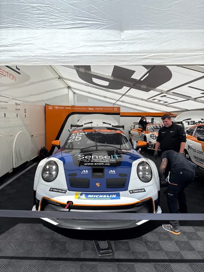 The scene is a garage pit stop at the 24 Hours of Spa-Francorchamps, an endurance sports car race held annually at the Circuit de Spa-Francorchamps in Stavelot, Belgium (latitude: 50.441, longitude: 5.969). A blue and orange Porsche 911 GT3 R race car with the number "25" is being serviced by two mechanics. The car is parked on a black and white checkered floor mat with the front facing the camera. The car has a "SenseIT" logo on the hood and the words "TAG Heuer" and "Michelin" on the front bumper. The mechanics are wearing black overalls and helmets. One mechanic is kneeling beside the car and another is standing next to the car on the driver's side. The garage is covered by a white tent with a large "U" shaped cutout in the center. In the background, a white and red race car is parked next to the car being serviced. The race car is facing the camera. A few people are standing near the cars.