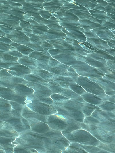 The image shows the surface of water in a pool or shallow body of water. The water is light blue-green and reflects sunlight in patterns of light and shadow. There are no people or objects visible in the water. There is no information about the location in the city of Germasogeia, Cyprus.