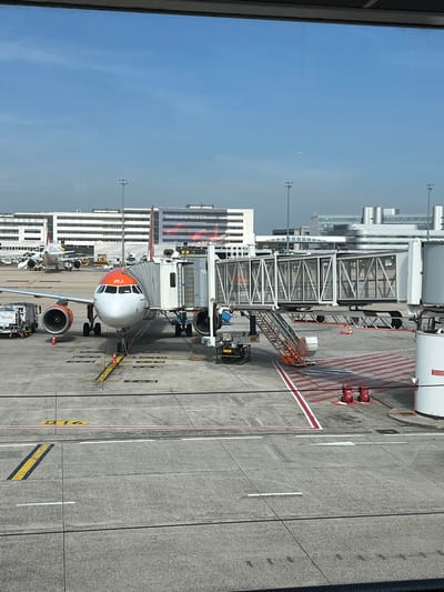 The image displays an airport apron scene at Charles de Gaulle Airport in Tremblay-en-France, France, observed from an elevated perspective through a window. The sky is clear and blue.
The central subject is a white and orange EasyJet Airbus A320 series aircraft, identifiable by its livery and the partial registration "INJ" on its nose, parked at gate D16. Its forward port door is connected to a multi-section passenger boarding bridge constructed with glass and metal panels. Below the boarding bridge, a mobile staircase with orange treads descends to the concrete apron. Ground support equipment is present: a ground power unit is connected near the aircraft's nose gear, and an air starter unit is positioned near the port engine. Orange traffic cones are placed around the aircraft's nose and wingtip. The concrete apron is marked with yellow taxiway lines, white service lines, red-and-white striped safety zones, and the prominent yellow gate designation "D16" within a red border. Two red wheeled fire extinguishers are situated on the apron near the jet bridge.
In the mid-ground, a second white aircraft, featuring a red tail, is visible further back on the apron, connected to a set of mobile boarding stairs. The background includes several large, multi-story airport terminal buildings of modern design. One building displays a large banner with the French text "S'ENVOLER EN TOUTE ÉLÉGANCE AIRF...". Utility poles with lighting fixtures are visible across the distant tarmac. No individuals are distinctly discernible engaged in specific actions within the frame.