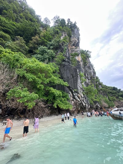 The image depicts a tropical beach scene on Ton Sai Bay, Koh Phi Phi Don, Thailand. A towering limestone cliff face, covered in lush green vegetation and bare rock, dominates the background. The sea in the foreground is a clear, shallow turquoise color, with small waves lapping at the sandy shore.

A group of approximately twenty tourists are visible on the beach and wading in the shallow water. They are dressed in a variety of casual beachwear, including swim trunks, shorts, and light shirts. Some are taking photographs with their phones, while others appear to be disembarking from or preparing to board a traditional Thai longtail boat anchored just offshore. The boat is white with a wooden hull and has Thai writing on its side.

On the far left of the image, a monkey is walking on the sandy shore. The beach itself is narrow, with the water meeting the dense vegetation that grows up the base of the cliff. The sky is overcast, suggesting a cloudy day, but the water remains bright and inviting. The overall atmosphere is one of a popular tourist destination experiencing a moderate level of activity.