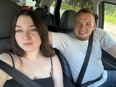 In Pesterevo, Russia, a young couple is seen inside a car, likely a sedan, with leather-like seats. The woman, with long dark hair and light eyes, is in the foreground on the left, looking towards the camera with a slight smile. She is wearing a black top and a seatbelt. To her right, the man, with short blonde hair and a beard, is also looking at the camera, smiling broadly. He is wearing a light-colored t-shirt and a seatbelt. The car's interior is visible, with the dark seats and the orange object on the back seat. Through the car windows, a glimpse of the Pesterevo environment is visible, with trees and greenery indicating an outdoor setting, possibly a road surrounded by nature. The overall impression is of a candid photograph taken during a car journey.