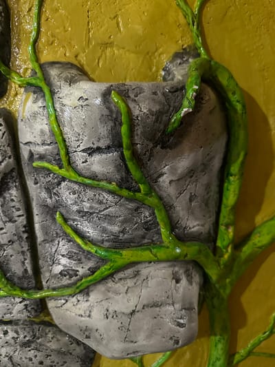 The image shows a close-up, detailed view of a textured relief artwork. The artwork depicts grey, rough-textured rocks, reminiscent of stone or concrete, with a vibrant green, vine-like element winding its way across and between them. The background is a solid, textured yellow surface. The green vine appears to be sculpted or molded with a glossy finish, showcasing some variations in thickness and branching patterns. The rocks also have a textured, slightly wet or glossy appearance, with visible striations and imperfections that give them a realistic feel. There are no people or indications of a specific location within the city of Kalininskaya, Russia, visible in this artwork. The focus is solely on the material and sculptural qualities of the piece.