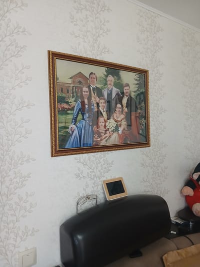 A family portrait is displayed in a ornate gold frame on a wall covered in light grey wallpaper with a subtle, repeating leaf-branch pattern. The painting depicts a group of eight people, likely a family, dressed in period clothing. In the foreground, a woman in a blue gown holds a bouquet of white flowers, seated next to a young girl in a light brown dress. Behind them are other figures, including two men in formal attire and several women. The background of the painting shows a brick building with columns, suggesting a historical or aristocratic setting.
Below the painting, on a black, curved surface, rests a pair of eyeglasses and a small digital device with a wooden frame and a black screen. To the right of these objects, a corner of a large, dark brown sofa is visible. Further to the right, a plush toy, possibly Mickey Mouse, with red and black coloring and pink ears, is partially visible on a small round table. The lighting in the room appears to be natural, coming from an unseen source to the left. The overall impression is of a domestic interior setting. This scene is located in Maslova Pristan', Russia.
