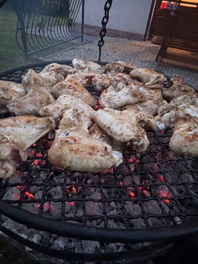 A close-up, overhead shot shows chicken wings being grilled on a round, black metal barbecue. The chicken pieces are irregularly shaped and have a light brown, cooked appearance with some darker seared spots. They are arranged loosely on the grill grate, with several wings overlapping. Beneath the grate, glowing red embers are visible, indicating the heat source. The background is out of focus, but suggests an outdoor setting, possibly a backyard or patio, with a glimpse of a fence and some greenery on the left, and a wooden structure and what appears to be a tabletop with bottles on the right. The lighting suggests it might be dusk or evening. This scene is located in Mordarka, Poland.